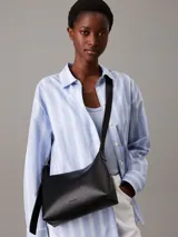 Bolso de hombro o cruzado de piel sintética negra, de forma rectangular y suave, con cierre superior de cremallera y correa ajustable con herrajes metálicos.