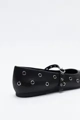 Zapato bajo tipo bailarina de color marrón oscuro, con puntera afilada y tira con hebilla sobre el empeine. El cuerpo del zapato está adornado con múltiples ojales metálicos plateados.
