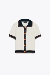 Chaqueta color blanco de punto con cuello solapa y manga corta. Tiene cierre frontal con botones color naranja y ribete en azul marino y naranja.