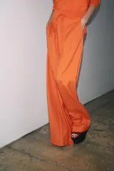 Pantalón ancho color naranja, de tiro medio y cintura con trabillas. Tiene bolsillos laterales y de vivo en espalda, pinzas en delantero, raya planchada y cierre frontal con cremallera, botón y ganchos metálicos.