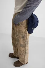 Pantalón de jean color beige con estampado abstracto floral en tonos marrones, de corte amplio, con bolsillos delanteros y traseros, y detalle de tira multifunción en la pierna. Cierre frontal con cremallera y botón.