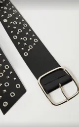 Cinto ancho de cuero sintético negro, con hebilla metálica plateada de forma cuadrada. La correa está decorada con múltiples tachas redondas y piramidales plateadas, además de ojales metálicos plateados a lo largo de toda su extensión.