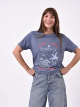 Remera blanca de algodón con cuello redondo y manga corta. Presenta un pequeño bordado en el pecho izquierdo con una figura que parece ser una persona acostada en una tabla de surf o flotador.