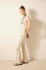 Pantalón de cuero ovino color crema con bordado de tachas metálicas y piedras en frente y espalda. De corte jeanero con calce flare, acceso delantero con botón metálico y cierre, bolsillos delanteros y traseros.