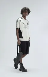 Chomba tejida de hilo color crudo con detalles en negro, de corte recto y manga corta. Presenta cuello tipo polo con rayas y bordado con el número 90 y la palabra 'Ravers'.