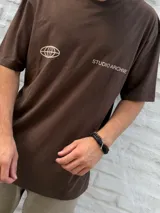 Remera marrón de manga corta, corte oversize, con un pequeño logo de un globo terráqueo estilizado y el texto "STUDIO ARCHIVE" estampado en el pecho.
