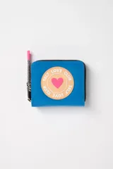 Billetera celeste con cierre de cremallera y tirador de color rosa. Tiene un parche redondo con borde color piel y un corazón rosa en el centro. El parche dice 'Self Love Club' alrededor del corazón.