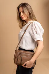 Bandolera de simil cuero color marrón topo, con textura ligeramente desgastada y detalles en nobuck. Presenta múltiples bolsillos frontales con cierres metálicos y tiradores de cuero. Correa ajustable para llevar al hombro o cruzada.