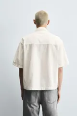 Camisa de denim de algodón, color beige claro, con corte relajado, cuello bowling y manga corta. Presenta cierre frontal con botones a presión y un pequeño bordado a tono en el pecho.