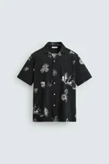 Camisa negra con estampado floral en tonos grises, de manga corta y cuello bowling. Confeccionada en tejido elástico que minimiza las arrugas. Cierre frontal con botones.