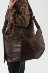 Cartera bandolera de cuero vacuno color chocolate, con diseño patchwork que combina cuero graneado, gamuza y cuero con grabado croco. Tiene correa ancha ajustable con tachas metálicas, forro interior de tela lisa, bolsillo con cierre y amplio espacio.
