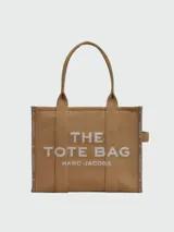 Bolso tipo tote de Marc Jacobs, modelo The Jacquard Large Tote Bag, confeccionado en tejido jacquard color tostado. Presenta el nombre de la marca y el modelo bordados en el frente en color blanco, doble asa superior y detalles de cintas laterales con inscripciones.