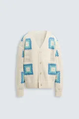 Cárdigan de punto color crudo con diseño de cuadrados de crochet en tonos celeste y azul. Tiene cuello en V, manga larga, bolsillos frontales y cierre con botones.