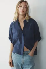 Camisa azul de cuello solapa y manga corta con dobladillo. Tiene dos bolsillos de parche en el pecho, bajo asimétrico con aberturas laterales y cierre frontal con botones ocultos.