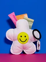 Monedero pequeño en forma de flor de tela acolchada color rosa pálido con carita sonriente bordada en amarillo. Incluye llavero con dije rosa.