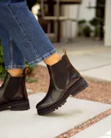 Bota tipo Chelsea de cuero negro con paneles elásticos laterales. Presenta una suela track de goma y un tirador de cuero marrón en la parte posterior.