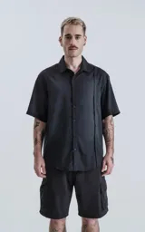 Camisa negra de manga corta con cuello clásico y cierre frontal con botones. Presenta una tira vertical cosida en el frente.