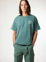 Camiseta de manga corta color verde azulado, corte holgado, con un pequeño estampado cuadrado con una 'X' en el pecho.