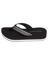 Ojotas de plataforma marca Tommy Hilfiger con tiras textiles negras que presentan un diseño de rayas blancas y el logo de la marca en relieve. La suela es de color negro con una línea blanca decorativa en el borde.