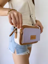 Bandolera rectangular de cuero color lila, con detalles en marrón y blanco. Tiene correa larga regulable, bolsillo frontal con cierre y etiqueta de cuero con logo de la marca.