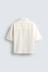 Camisa de denim de algodón, color beige claro, con corte relajado, cuello bowling y manga corta. Presenta cierre frontal con botones a presión y un pequeño bordado a tono en el pecho.