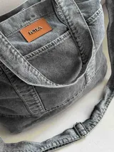 Bolso grande tipo totebag de jean azul, con asas de mano y asa larga para colgar. Se cierra mediante 1 botón de metal a presión y contiene 3 bolsillos exteriores.