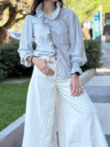 Camisa celeste de lino metalizado con volados en el cuello y en el frente. Tiene mangas largas con puños elásticos y botones escondidos.