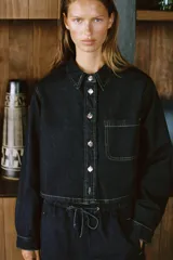 Camisa corta de denim celeste, con cuello solapa, manga larga, bolsillo delantero de plastrón y cierre frontal con botones metálicos.
