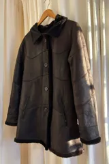 Abrigo gamulán marrón con cuello, solapa, puños y borde frontal de piel sintética blanca. Tiene cierre con botones y bolsillos laterales.