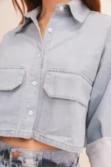Camisa de denim azul con cuello clásico, cierre frontal con botones blancos, mangas largas con puños abotonados y dos bolsillos cargo con solapa en el pecho.