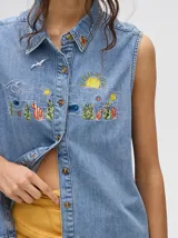 Camisa sin mangas de denim liviano color camel, con cuello camisero y botones frontales. Presenta bordados temáticos marinos en el pecho, incluyendo un sol, olas, una gaviota y vida marina.