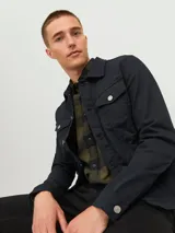 Chaqueta de denim negra con cuello clásico, cierre frontal con botones metálicos plateados, bolsillos con solapa abotonada en el pecho y mangas largas con puños abotonados.