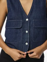 Chaleco denim azul oscuro, con cuello en V, cierre frontal con botones y bolsillos de parche.