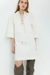 Vestido corto de denim color blanco, con cuello camisero, mangas cortas, dos bolsillos frontales y ajuste con cordones cruzados en el escote.