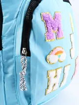 Mochila celeste con ribetes negros, asa superior y correas acolchadas ajustables. Decorada con parches de letras y símbolos en colores brillantes y texturas variadas.