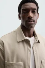 Sobrecamisa negra de tejido con elasticidad, cuello solapa y manga corta. Tiene dos bolsillos de plastrón en el pecho y cierre frontal con botones a presión.