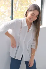 Camisa blanca de manga larga con puños, cuello camisero y cierre frontal con botones ocultos. Detalles de entredós a tono.