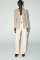 Blazer color beige, sin solapa, de manga larga con bolsillos de vivo en el delantero y cierre frontal con gancho metálico.