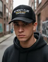 Gorra trucker negra con frente rígido y rejilla trasera. Bordado frontal con la frase "No Mercy Club" en blanco y amarillo.