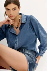 Camisa celeste de tencel con calce recto, manga larga amplia con frunces en los hombros y cuello camisero. Mangas terminadas en puño con botón y ojal. Cartera delantera con botones metálicos con el logo de la marca.