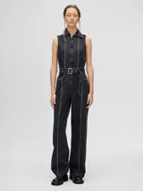 Mono largo de denim negro con cuello camisero, cierre frontal con cremallera, sin mangas, corte recto y cinturón ajustable. Decorado con tachas plateadas en cuello, cierre, cintura y costuras laterales.