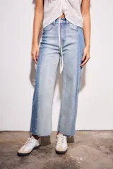 Jean de tiro medio, corte recto y largo cropped, confeccionado en denim de algodón con elastano. Presenta un efecto de lavado bicolor o combinado, con paneles de diferente tono de azul claro, y costuras verticales marcadas en el frente. El ruedo es deshilachado.