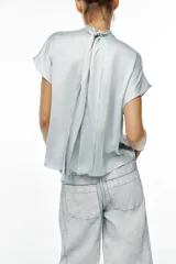 Blusa color crudo de tejido satinado, con cuello subido, manga sisa y cierre en la espalda con lazada y botones.