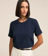 Blusa blanca de poliéster con cuello redondo, mangas cortas, cierre trasero en forma de gota, pequeñas aberturas laterales y acabados en pespuntes en las mangas y el cuello.