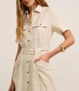 Vestido midi color beige confeccionado en sarga, con cuello camisero, manga corta, abotonado frontal, bolsillos utilitarios en el pecho y lazo en la cintura.