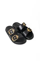 Sandalias planas tipo 'slide' de cuero con dos tiras anchas ajustables mediante hebillas doradas. La plantilla es negra con el logo 'MACU' en dorado. Se muestran en color negro y color bordó (vino tinto) charol.