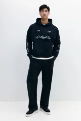 Sudadera tipo canguro de algodón pesado con efecto lavado oscuro, corte oversized y hombros caídos. Presenta estampado frontal en blanco con texto cursivo "Le Strade Di MMXXVI", dos banderas a cuadros, texto pequeño en la parte superior derecha y texto vertical en la manga izquierda. La manga derecha tiene un patrón de estrellas delineadas.