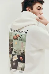 Buzo canguro color crema con capucha cruzada de doble capa, hombros caídos, bolsillo tipo canguro, puños y bajo acanalados. Estampado frontal con flores y texto "Beautanical". Corte holgado.