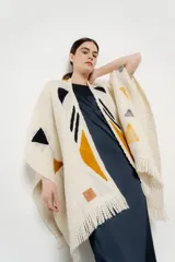 Ruana de tejido grueso color crema con flecos en el borde inferior y diseño geométrico en ocre, negro y gris.