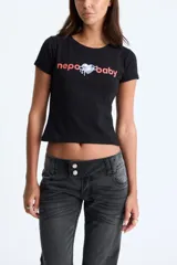 Remera negra de manga corta y corte ajustado, con estampado frontal que dice "nepo baby" en letras rojas y un corazón plateado con efecto de goteo en el centro.